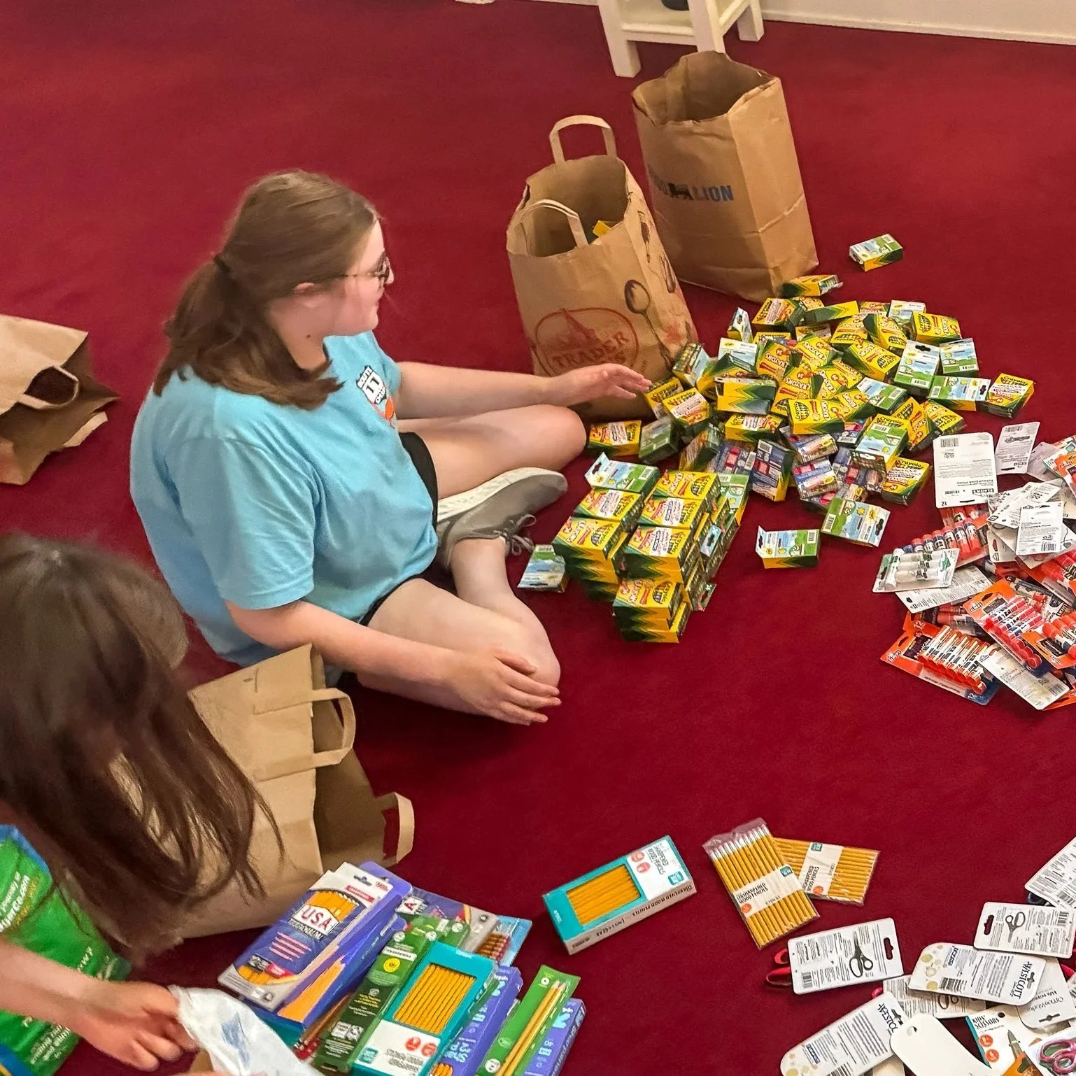Several people sitting on the floor surrounded by school supplies like notebooks, scissors, and packs of pencils, surrounded by paper bags and larger paper grocery bags.