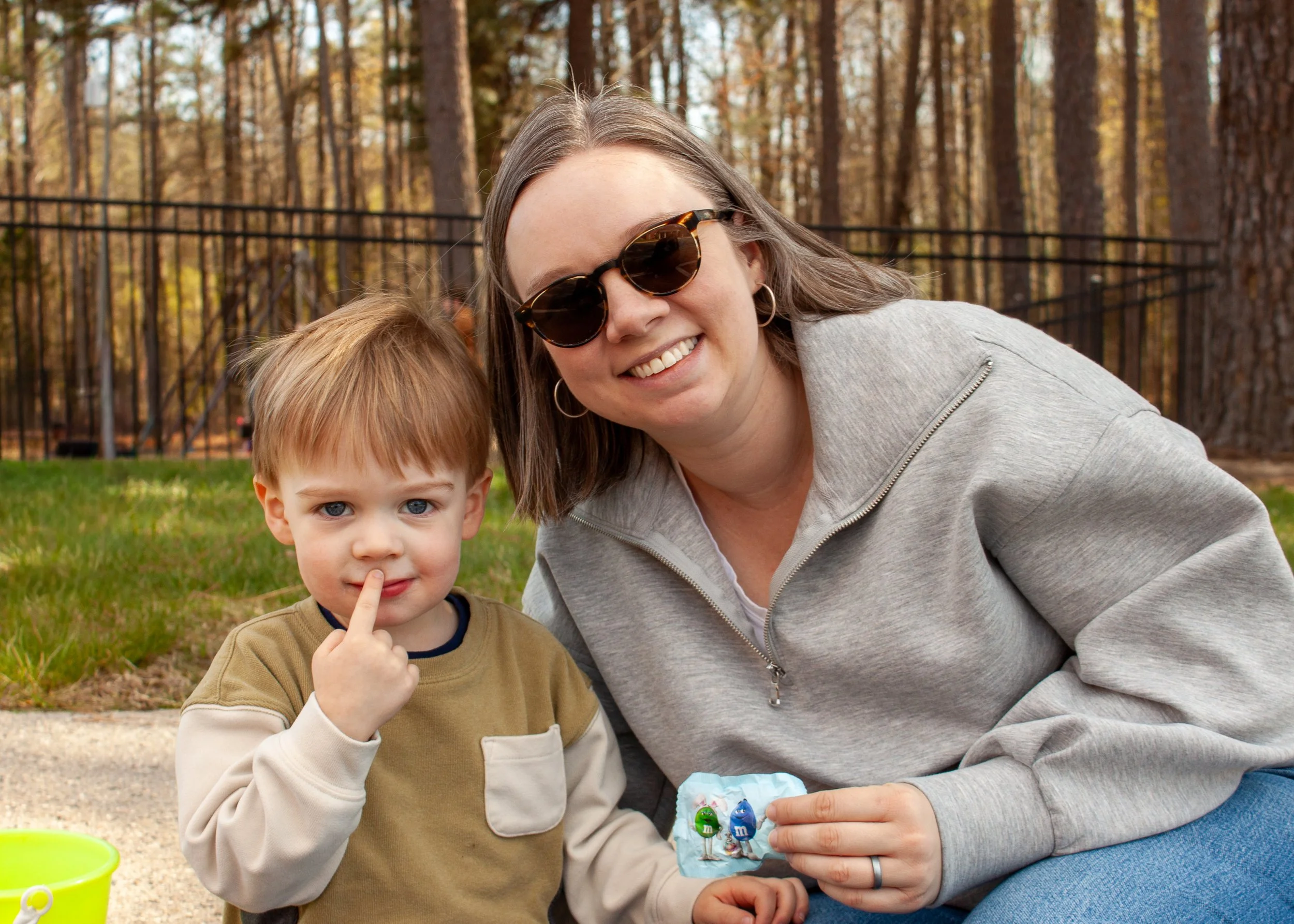 A woman wearing sunglasses and hoop earrings smiling with a young boy who has light brown hair and blue eyes, sitting outdoors near a wooded area with a black metal fence in the background.
