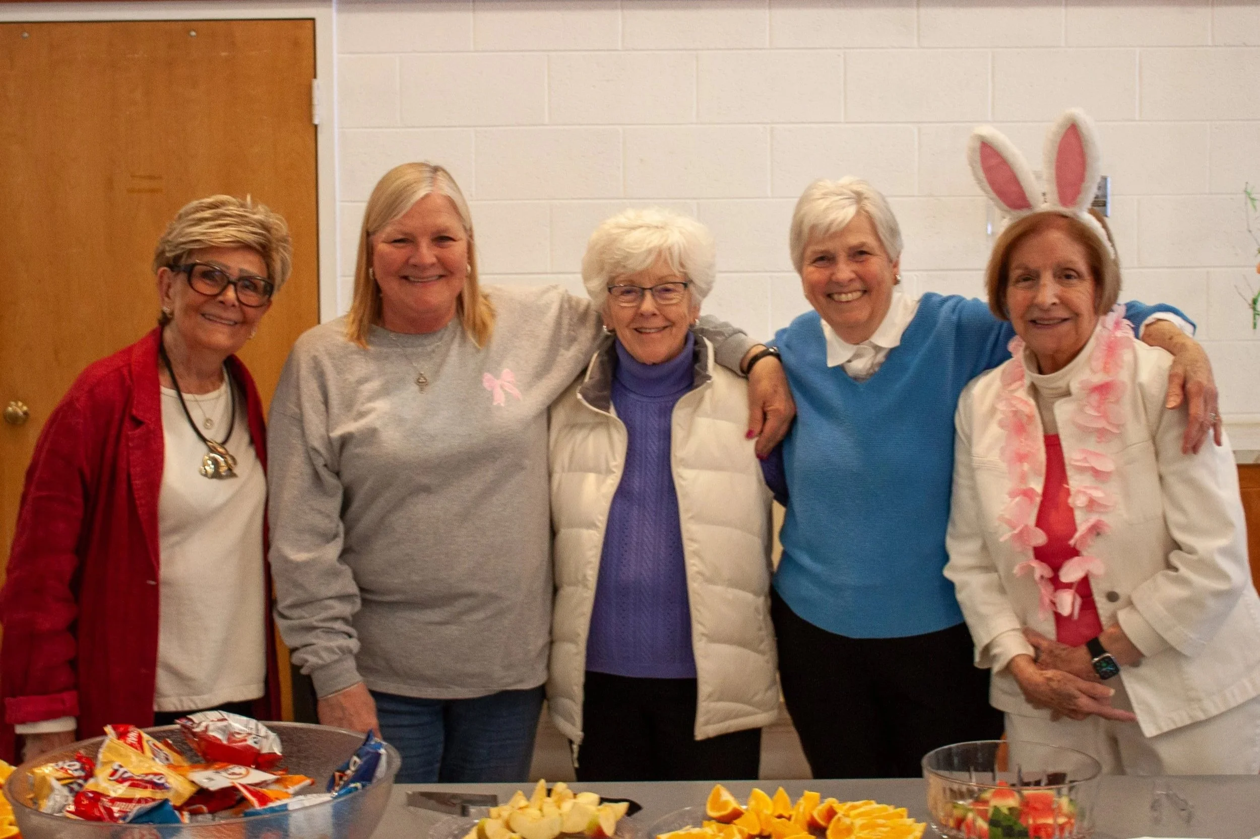 Five women standing together, smiling, with snacks and fruit on a table in front of them, in a room with white brick walls and a wooden door.