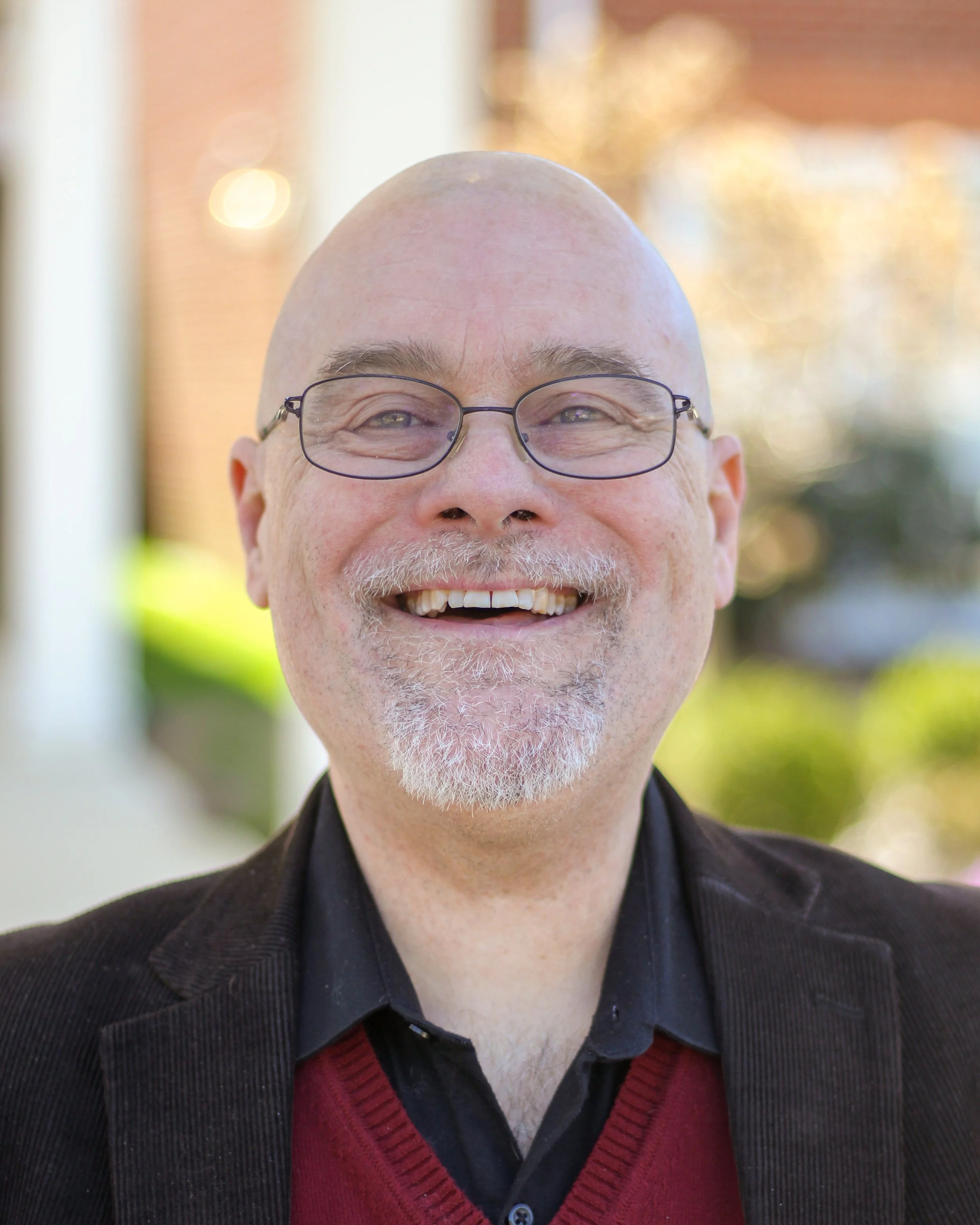 Close-up of a smiling man with glasses, bald head, and gray beard, wearing a black jacket and red sweater, outdoors with blurred background of lights and trees.