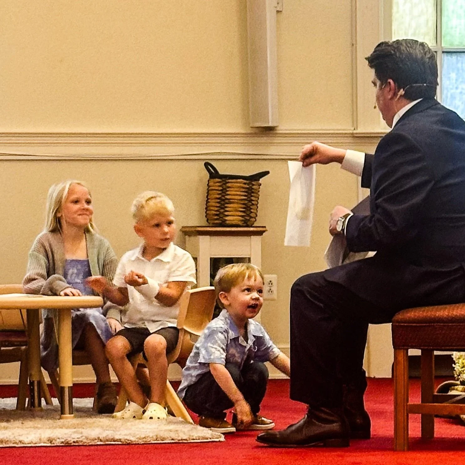 A man dressed in a suit is performing a puppet show for four children sitting on the floor and chairs in a room with a beige wall and window, wooden furniture, and a basket on a small table.
