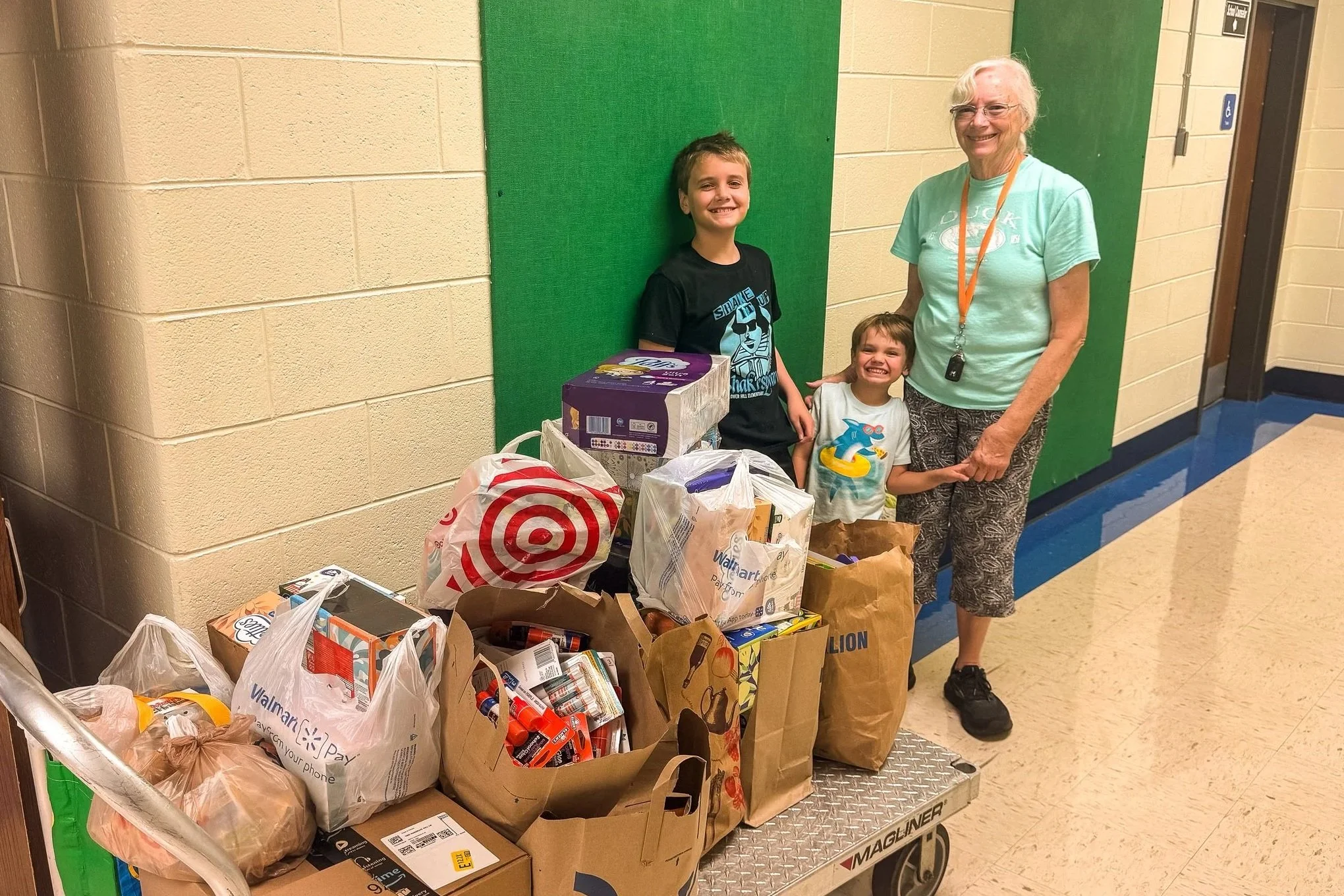 A smiling boy, a young girl, and an older woman standing behind a cart filled with grocery bags, in a hallway with beige and green walls, next to an elevator.