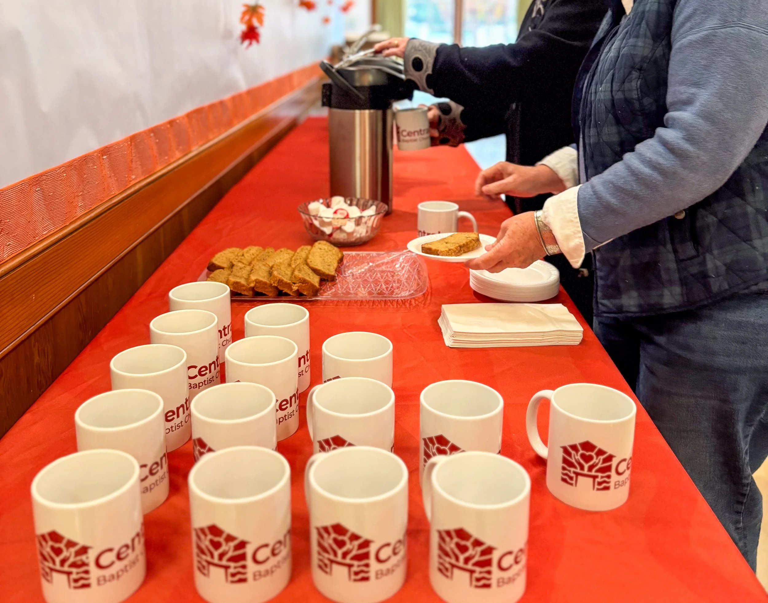 People serving coffee and baked goods on a red tablecloth, with cups displaying 'Centennial Baptist' and a gingerbread cookie on a plate.