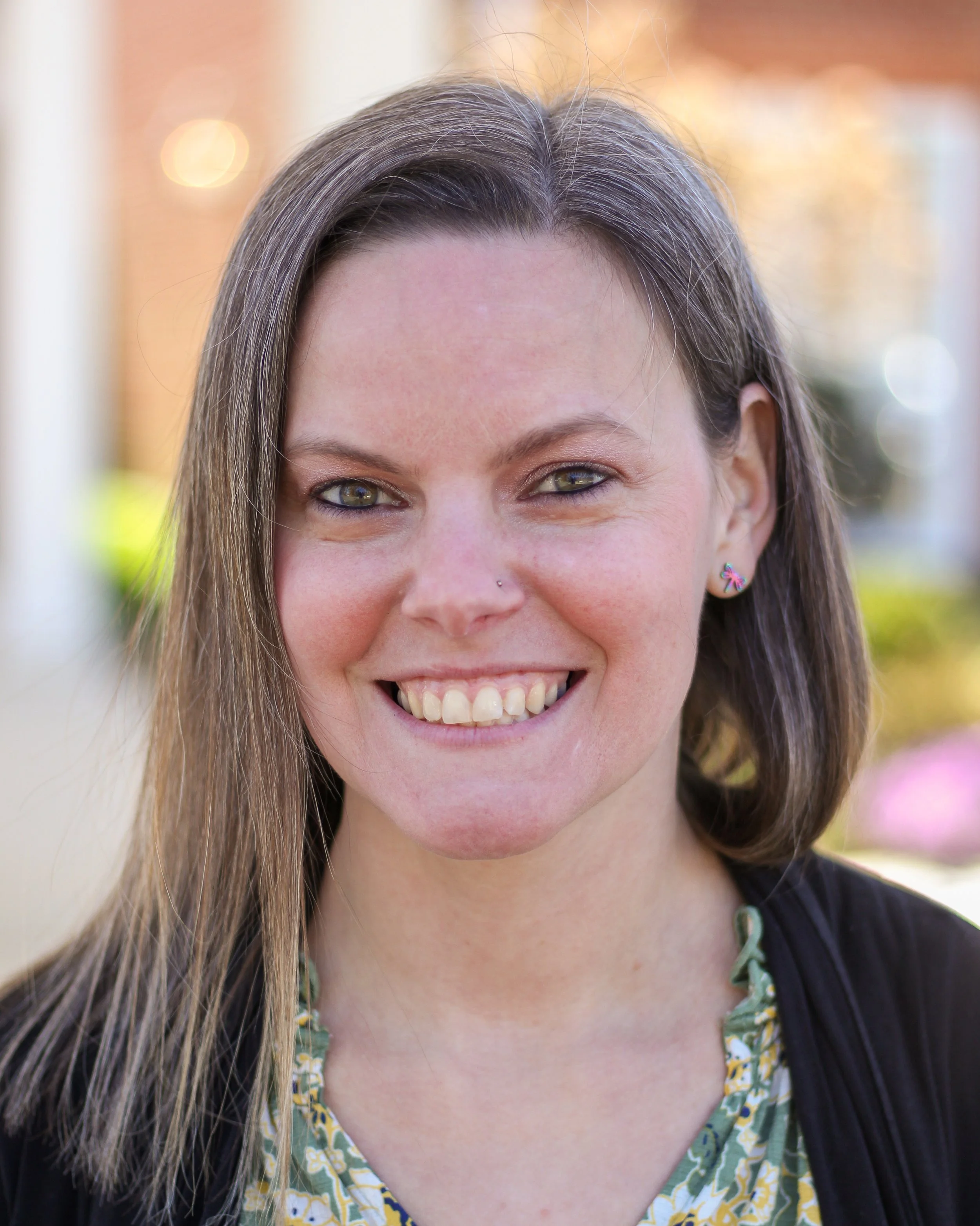 Close-up of a smiling woman with long, brown hair, green eyes, and a small nose piercing. She is wearing a floral shirt and a dark vest, standing outdoors with blurred trees and sunlight in the background.
