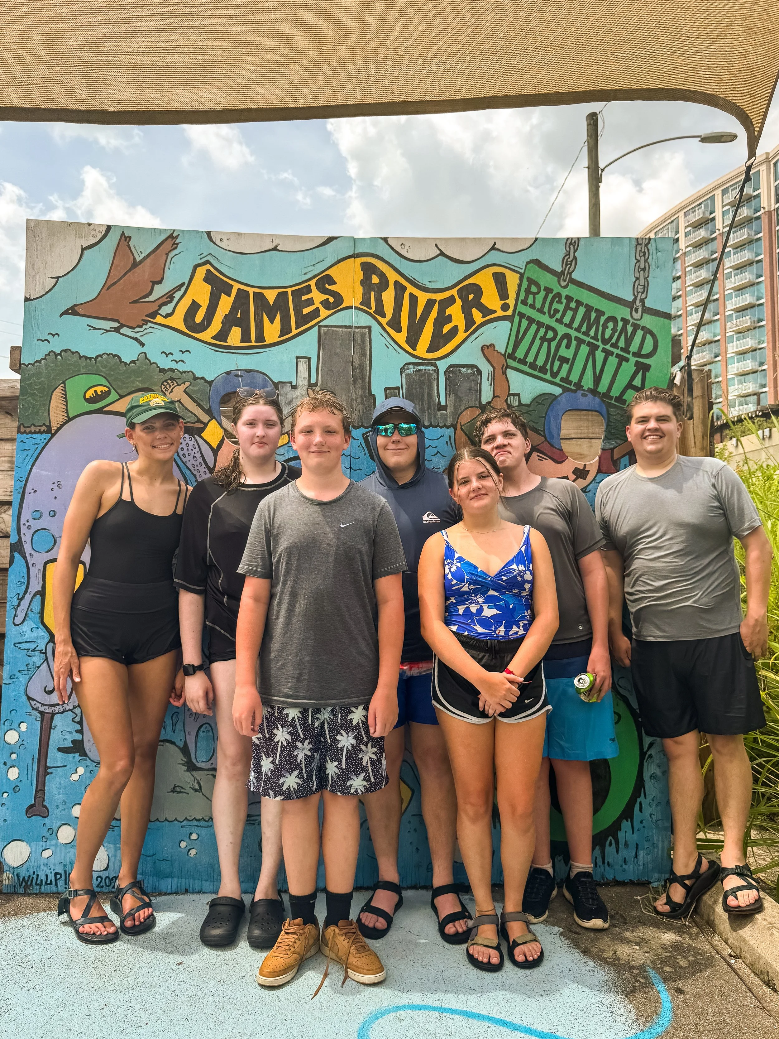 Group of seven teenagers standing in front of a colorful mural that says "James River! Richmond Virginia" during daytime, with one person holding a green water bottle, some wearing sunglasses and summer clothing, in an outdoor setting with a building and power lines in the background.