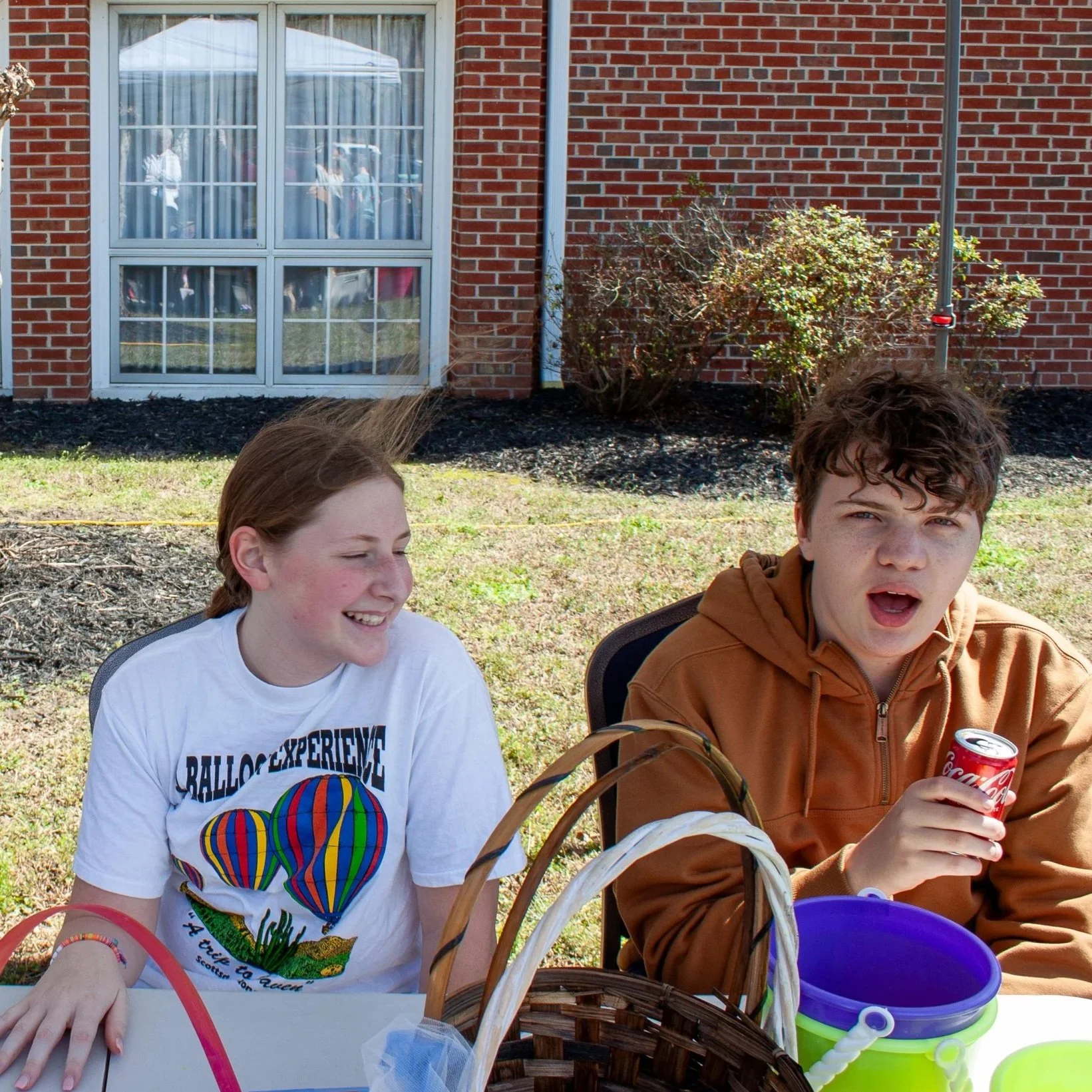 Two teenagers sitting outdoors near a brick building with a window, one girl with red hair smiling and a boy with curly brown hair holding a Coca-Cola can, on a sunny day.