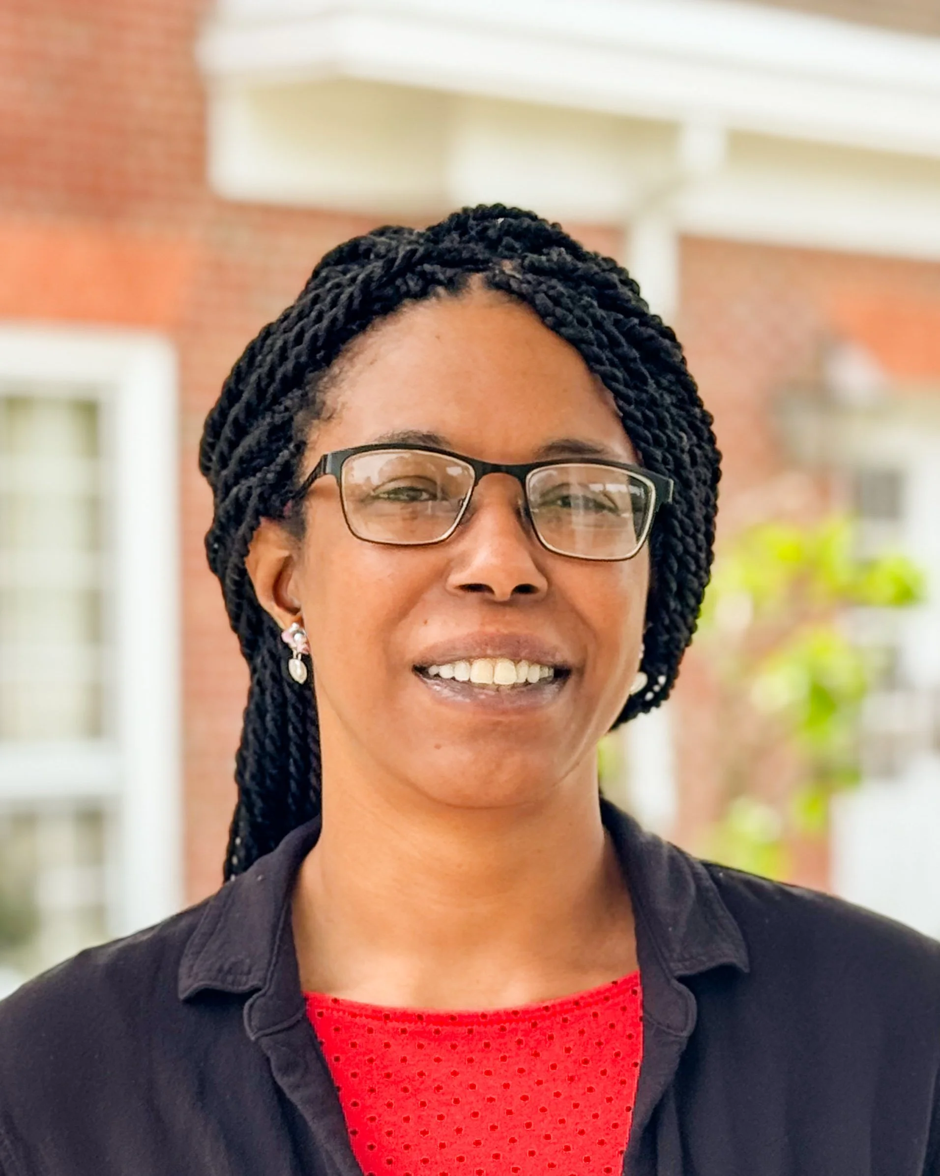 Close-up of a smiling woman with braided hair, glasses, earrings, wearing a red shirt and a black jacket, standing outdoors with a brick building background.