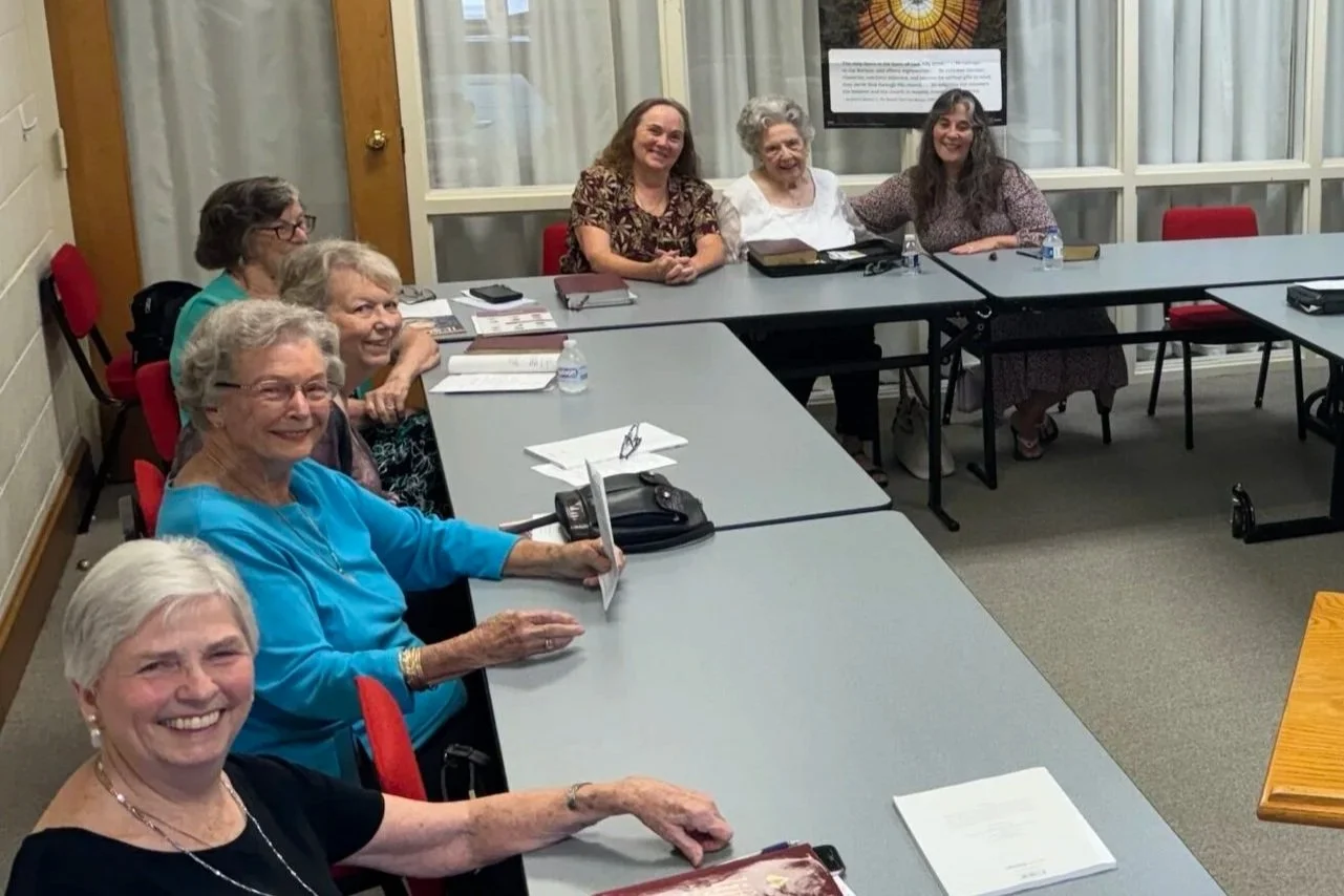 Six elderly women sitting around a U-shaped conference table in a meeting room, smiling. Books, notebooks, water bottles, and papers are on the table.