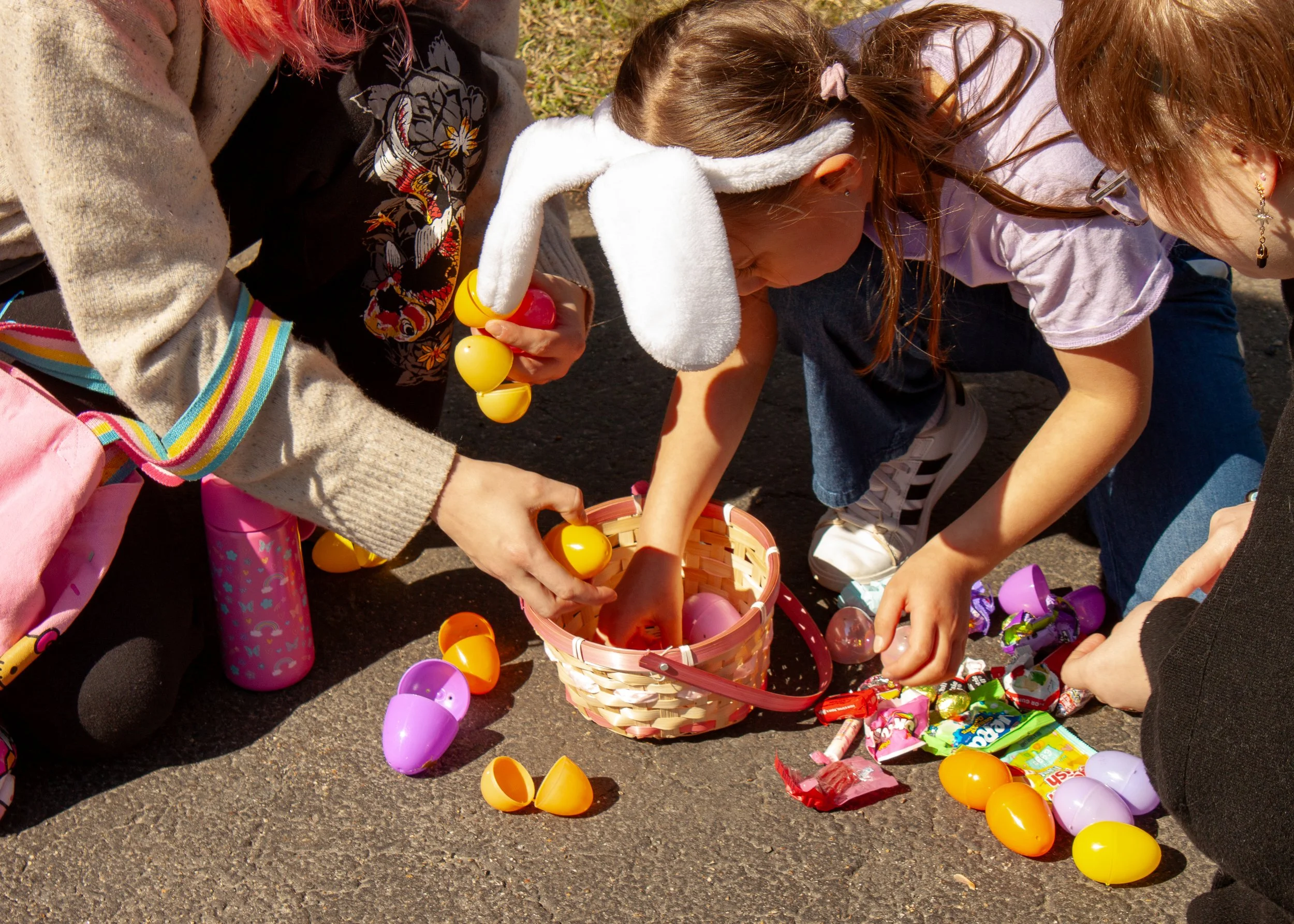 Children participating in an egg hunt, collecting plastic eggs and candy from the ground on a sunny day.