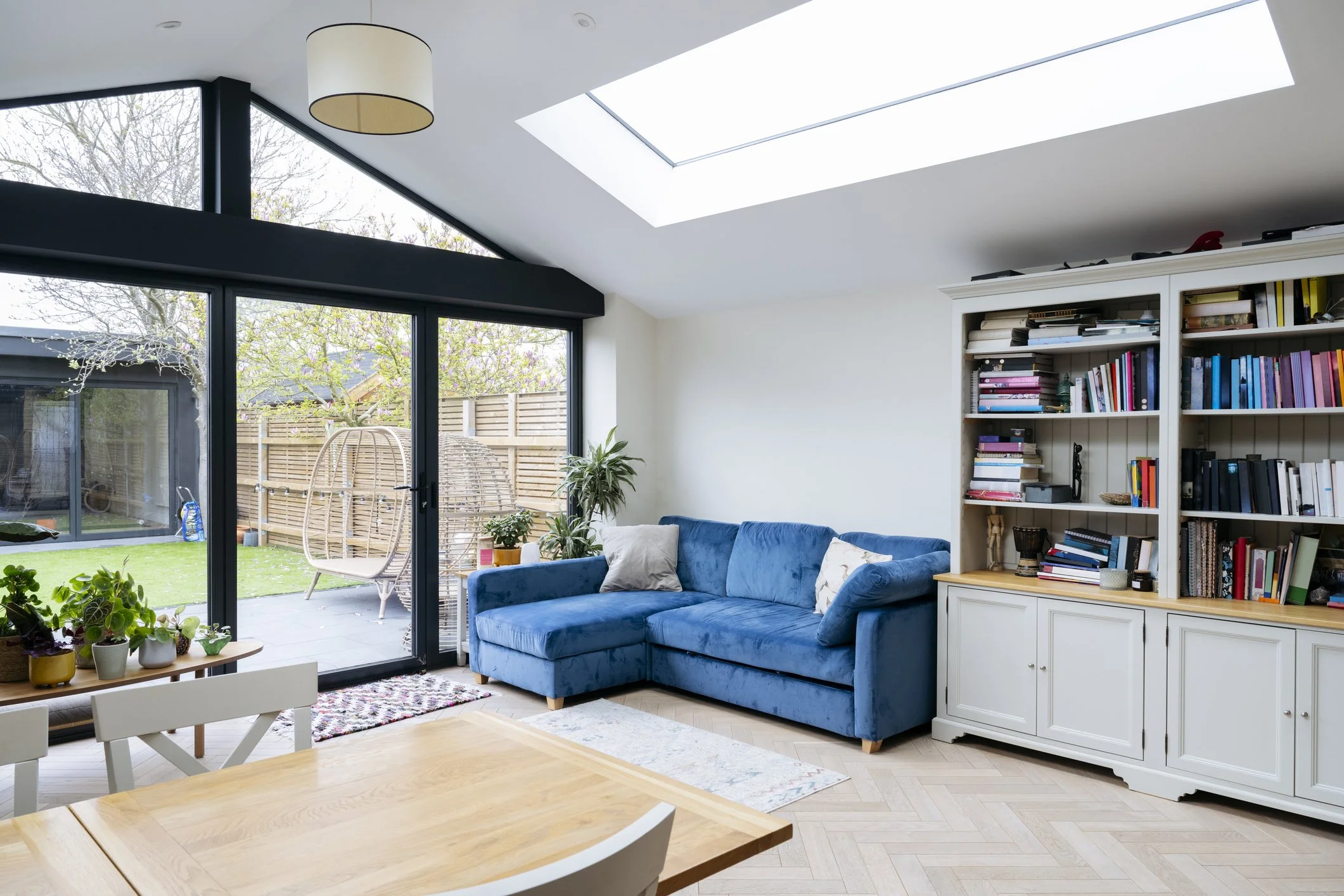 A bright blue couch sits against a wall made entirely of windows, letting in lots of natural light to this custom addition. Plants, a light wooden table, and the features of home fill out the image.