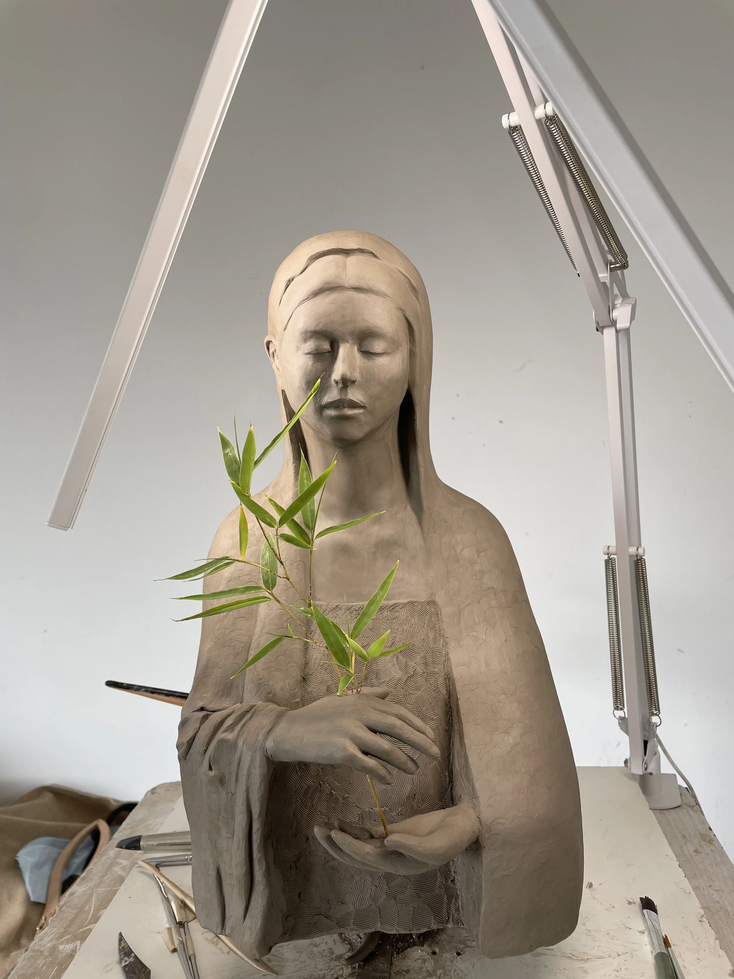Clay sculpture of a woman with long hair holding a small bamboo plant.