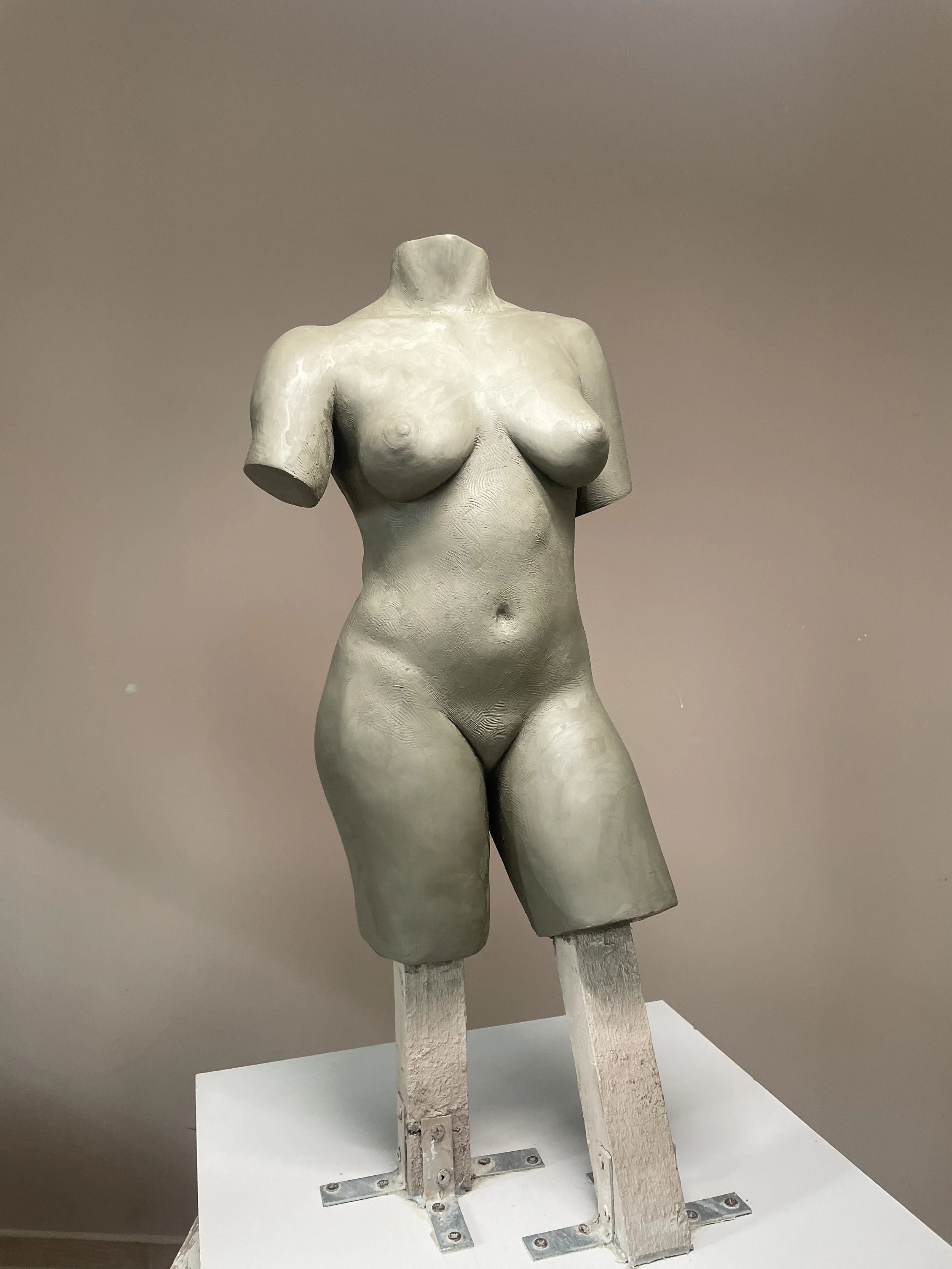 Unfinished sculpture of a female torso with no head or arms, mounted on a stand, in a neutral-colored room.