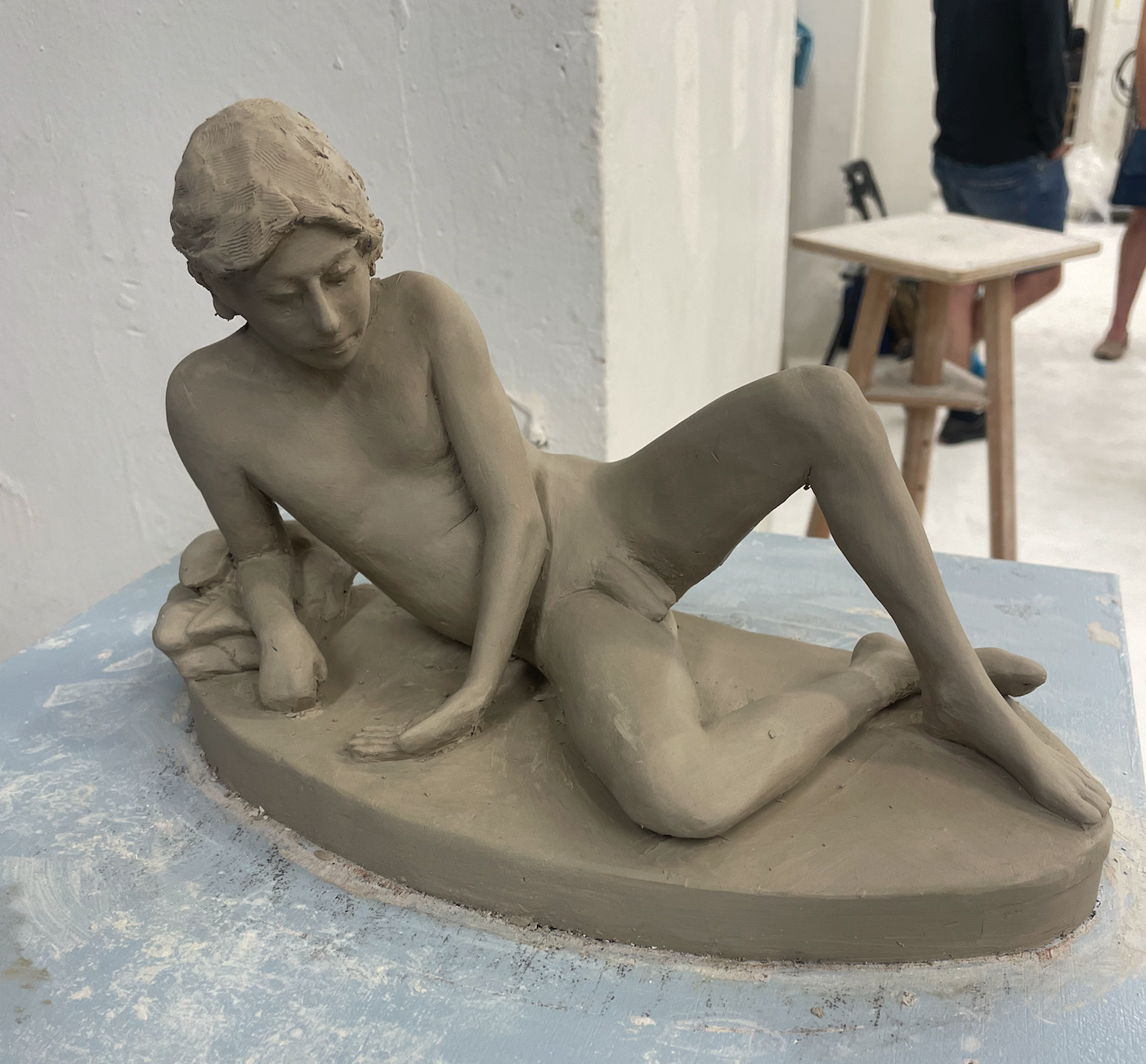 A clay sculpture of a young man lying on his side with one arm propping him up and the other hand on the ground, with detailed facial features and hair, placed on a worktable in an art studio.