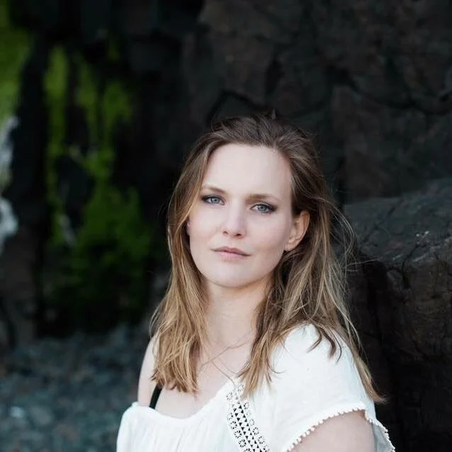 A woman with light brown hair and blue eyes standing outdoors near a rocky background with greenery.