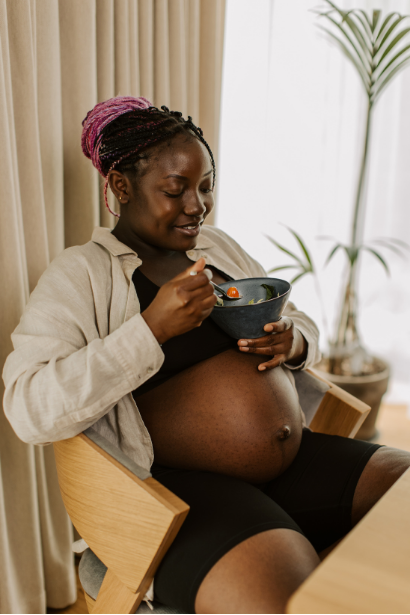Pregnant woman with pink braided hair eating from a bowl while sitting in a room with beige curtains and potted plants.