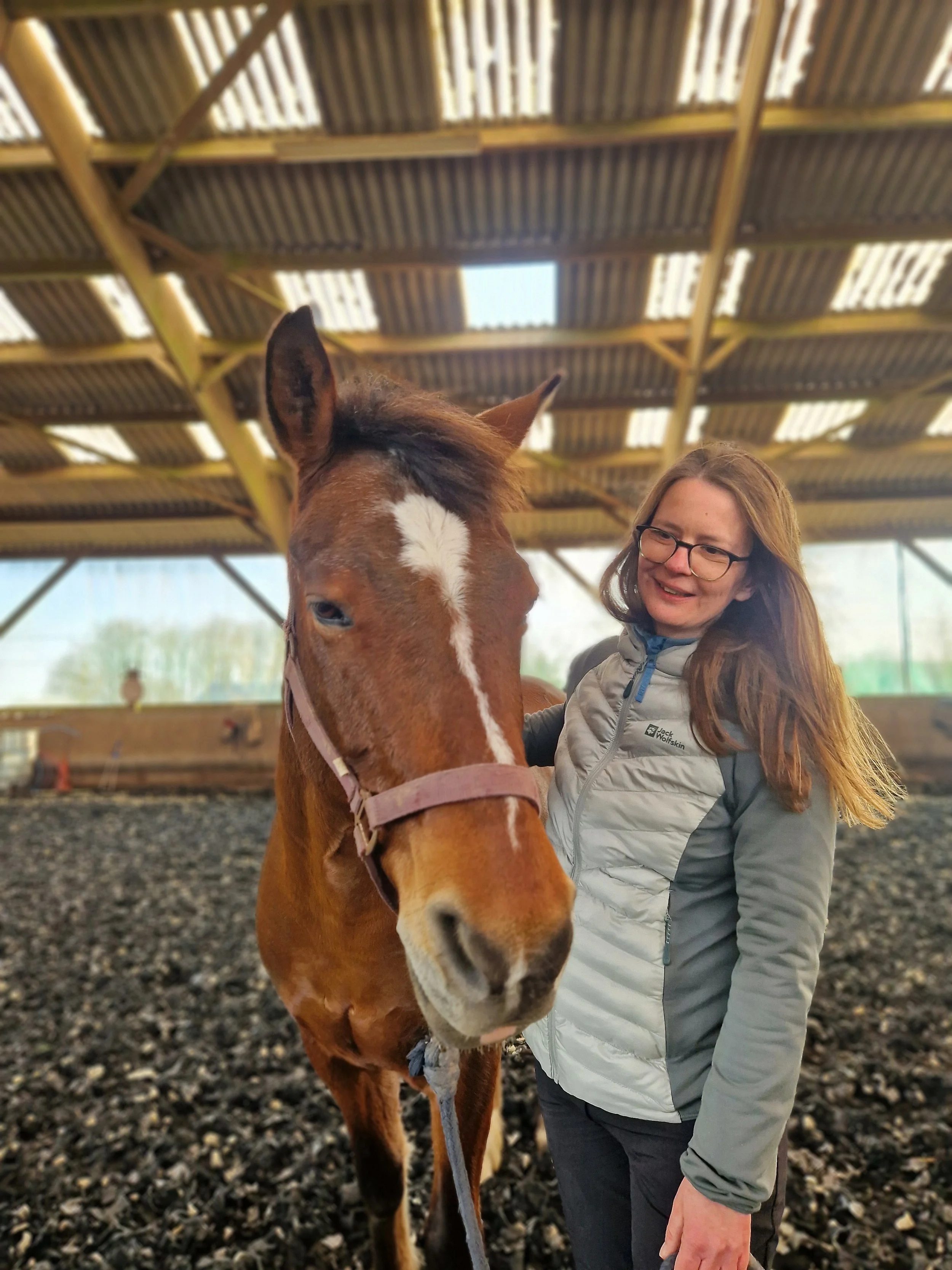 Junge Frau mit Brille und grauer Jacke steht im Reitstall, lächelt und streichelt ein braunes Pony unter einem holzvertäfelten Dach.