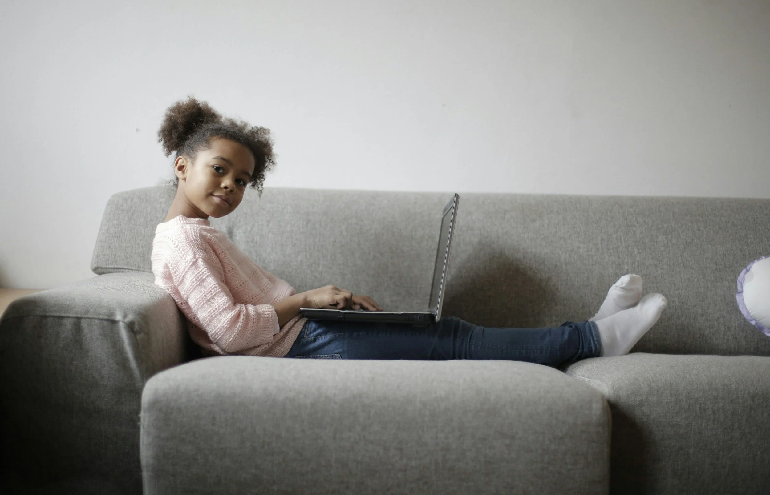 A young girl with curly hair, wearing a pink sweater and white socks, lies on a gray couch using a laptop.