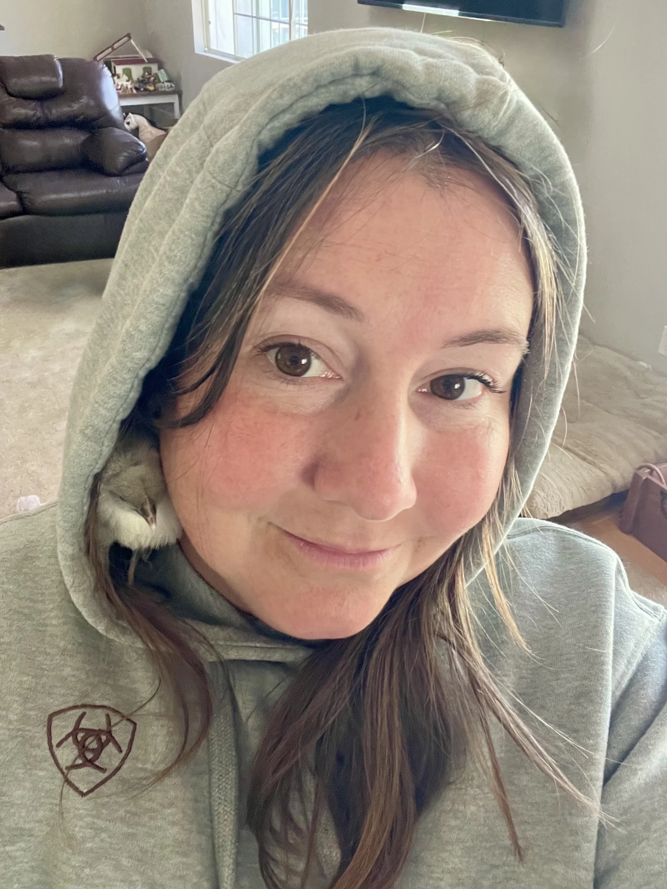 A woman with brown hair and brown eyes smiling while wearing a green hoodie with a Ariat logo on the chest. She is indoors with a chick peeking out from inside her hoodie.