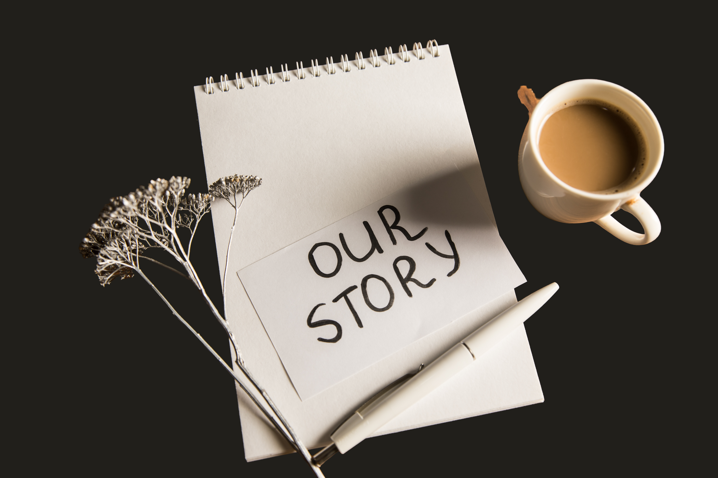 An open notebook with a handwritten note saying 'OUR STORY', a pen, a dried flower, and a cup of coffee on a black surface