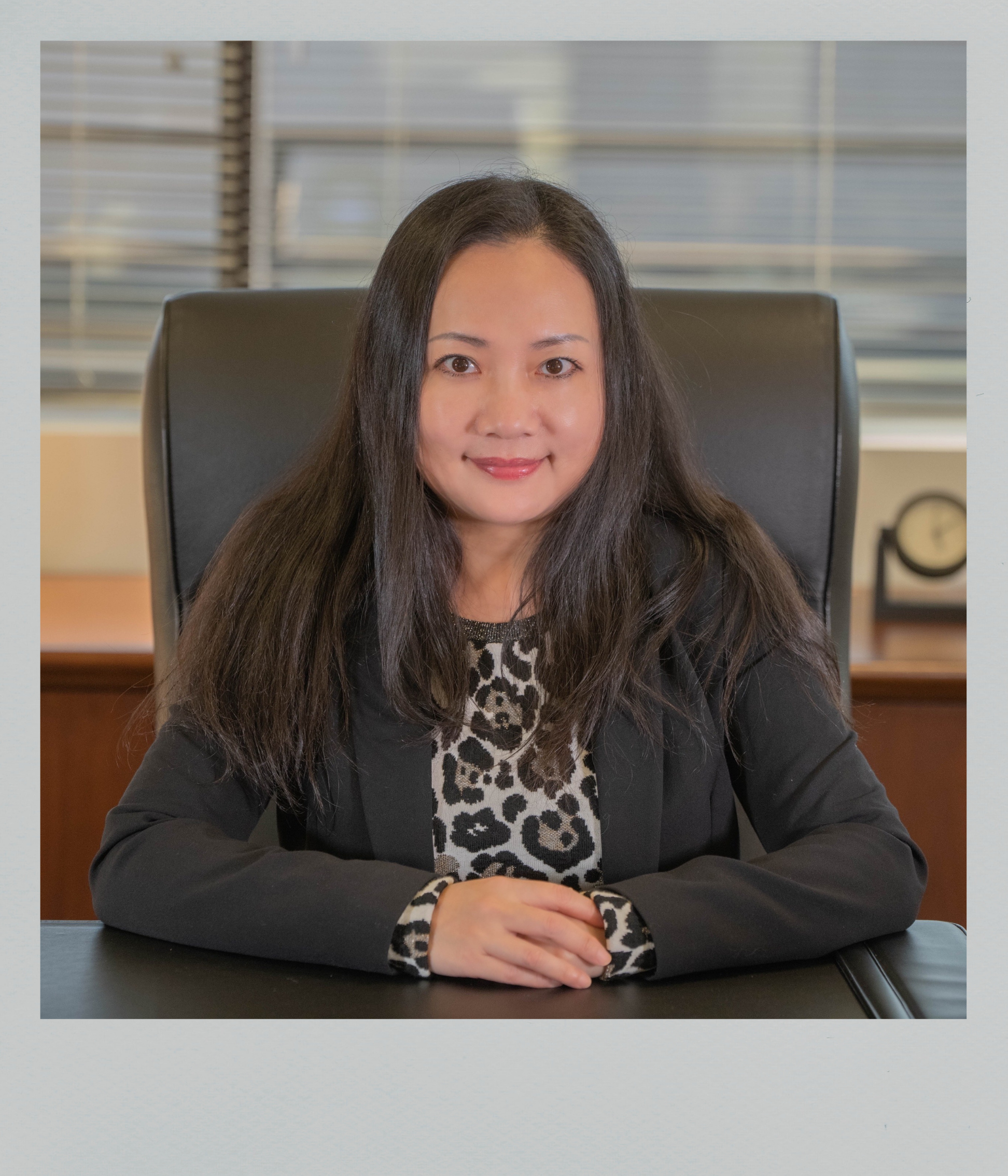 A woman with long black hair smiling while sitting at a desk in an office, wearing a black blazer over a leopard print top.