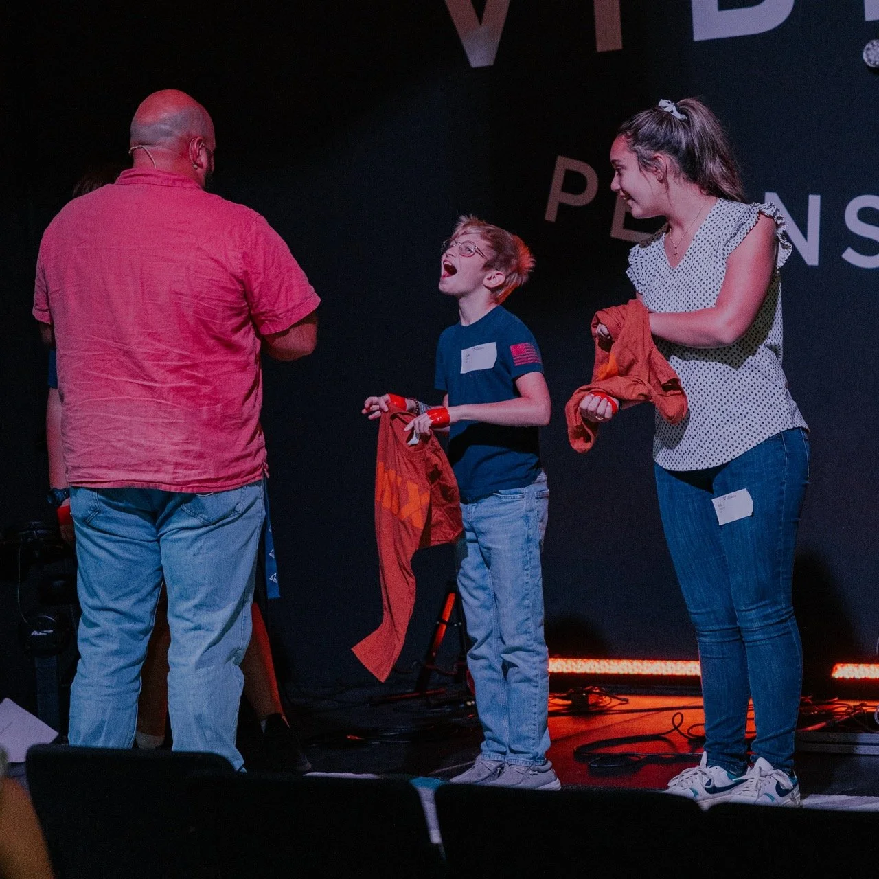 A young boy with glasses and a woman are on a stage, smiling and holding orange shirts. A man with a bald head, wearing a red shirt, is talking to them, possibly during an award ceremony or event.