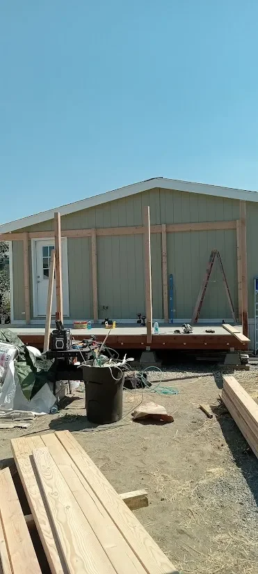 A house under construction with exposed wooden framing and a deck being built, various construction tools and materials are scattered around on a dirt surface under a clear blue sky.