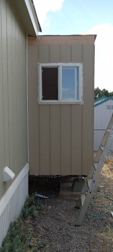 Small beige structure with a window, attached to a green house, with a ladder leaning against it.