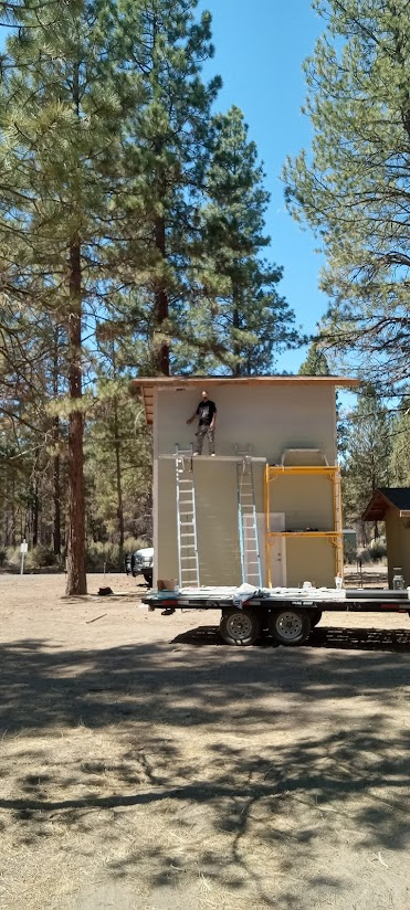 A person working on a small construction project on top of a partially built shed, with ladders leaning against the shed, in a wooded outdoor area.