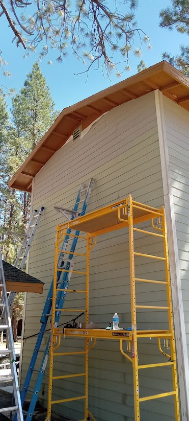 A house with light-colored siding and a sloped roof under a clear blue sky, with tall pine trees in the background. There are three scaffolding units and two ladders set up against the house, indicating ongoing maintenance or construction work.