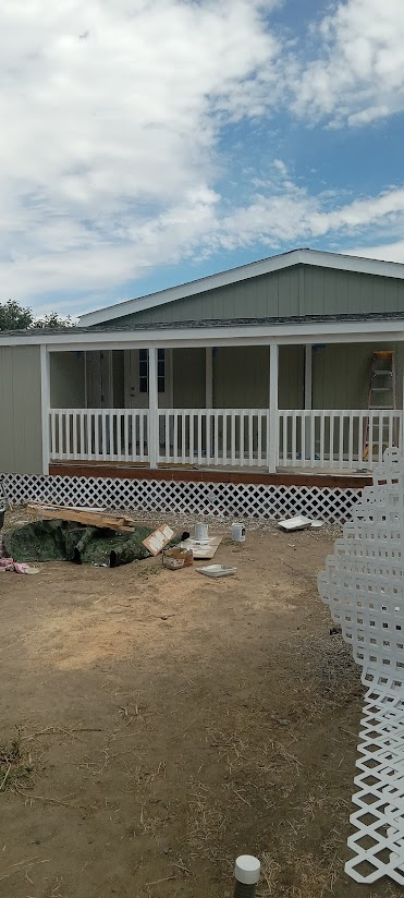 A house with a raised porch under construction, surrounded by construction tools and materials on bare ground, with a partly cloudy sky overhead.