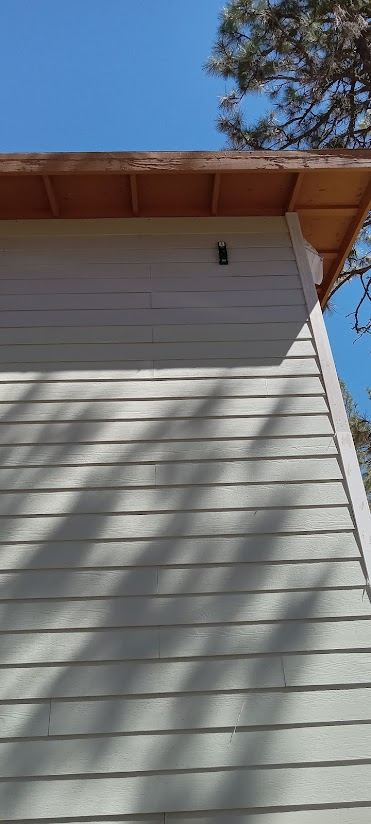 Close-up of the corner of a house with white horizontal siding under a wooden roof edge, with a clear blue sky and a tree in the background.