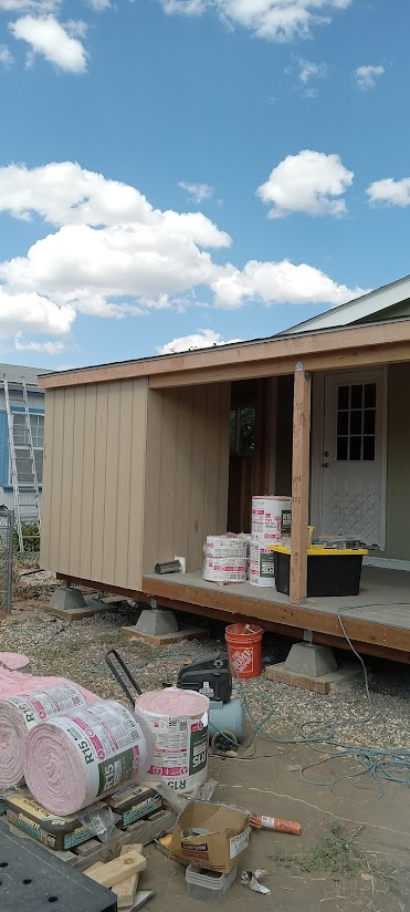 Construction site of a house with unfinished porch, building materials, tools, and an orange bucket in front of a partly cloudy sky.