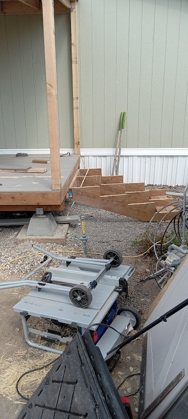 Wooden staircase under construction next to a partially built porch with light green siding, with construction tools and equipment on the ground.