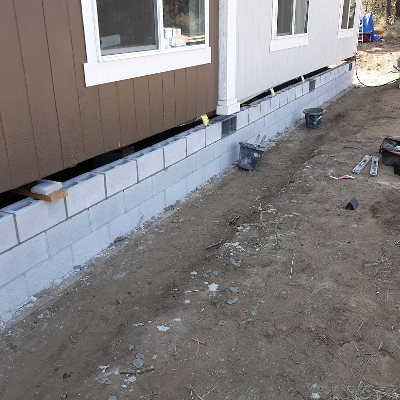 Construction site with cinder blocks forming a foundation along the base of a house, with tools and buckets nearby.