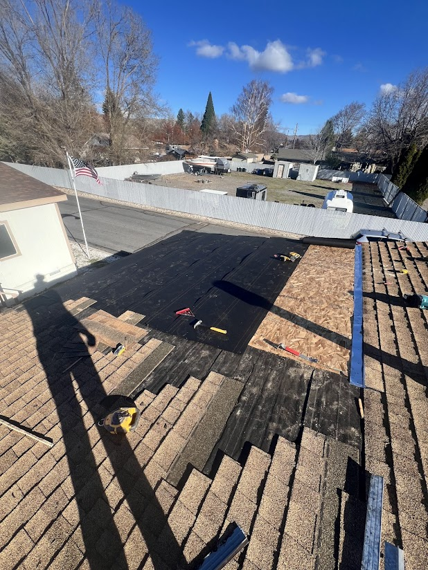 A residential roof is under repair with new black roofing material being installed. Tools and materials are scattered on the roof, and a person’s shadow is visible in the foreground. The background shows a neighborhood yard with trees, trailers, and 