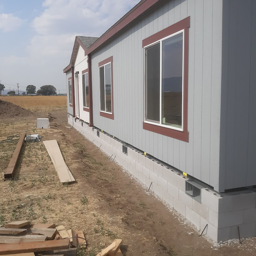 Exterior view of a house under construction with gray siding, brown window frames, and a concrete block foundation. Building materials and tools are scattered on the ground nearby.
