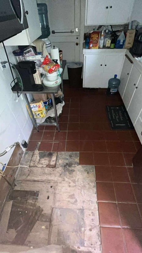 Small kitchen with white cabinets, a black water dispenser, a cluttered counter with food boxes and utensils, and a floor transition with missing tiles and exposed subfloor.