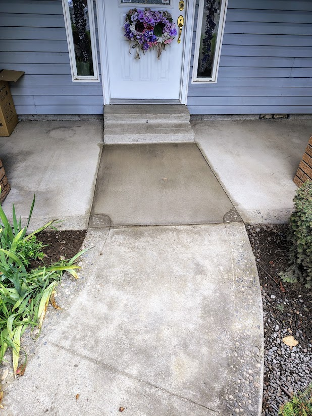 Concrete walkway leading to front door with a floral wreath, flanked by gray siding and small garden beds.