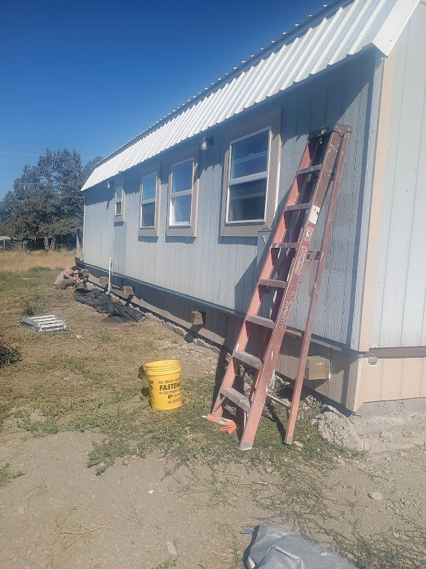 A house with blue siding and four windows under a white metal roof, a ladder leaning against the side, a yellow bucket on the ground, and construction materials nearby.