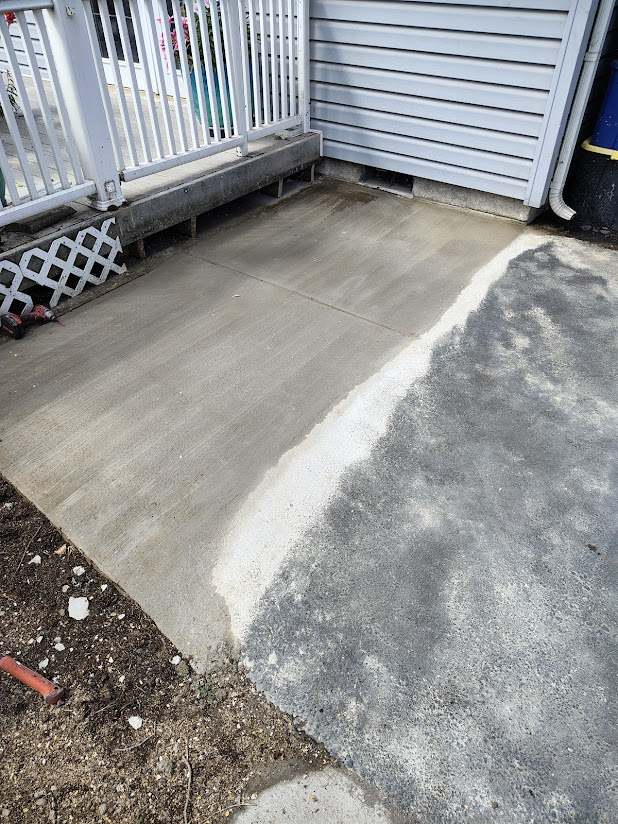 A freshly poured concrete patio at the corner of a house with a metal railing, a lattice panel, and some construction tools visible at the edge.