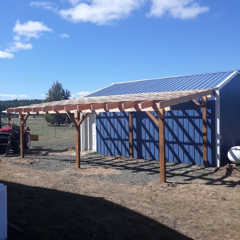 Wooden structure in front of a blue metal building, possibly a carport or shelter, with a tractor parked nearby under clear skies.