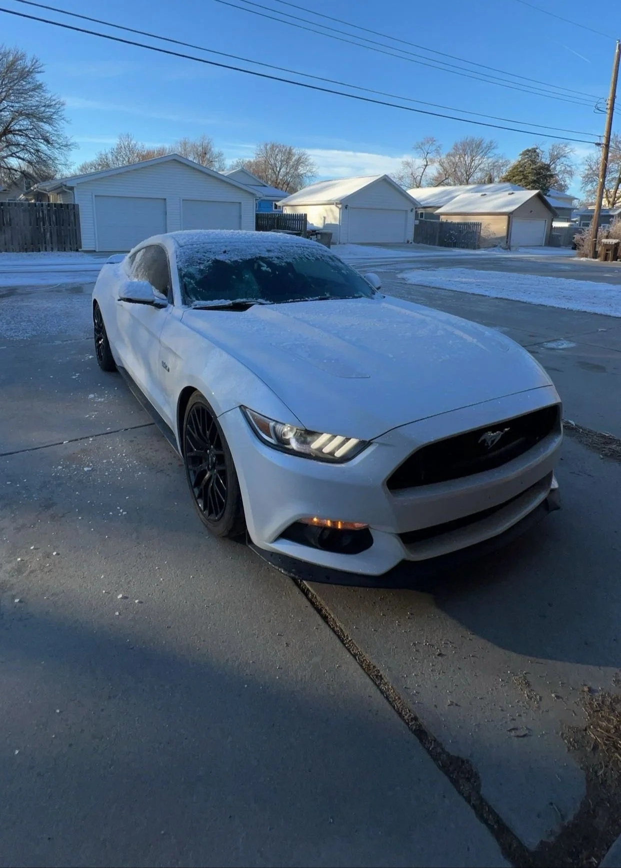 A white Ford Mustang parked on a driveway in a snowy suburban neighborhood, with snow on its roof and windshield, under a clear blue sky.