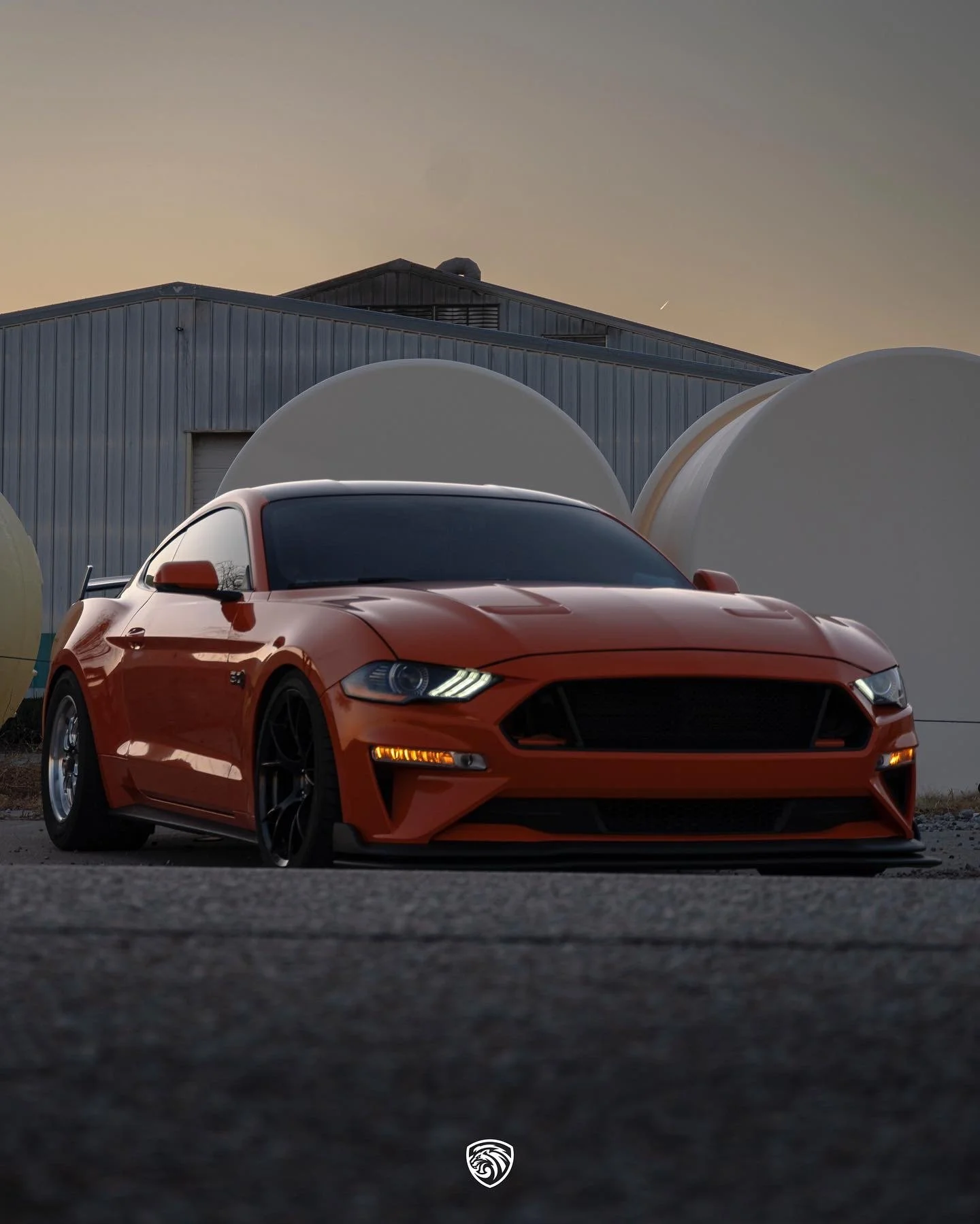 A red sports car parked outdoors during sunset with a warehouse and large white storage tanks in the background.