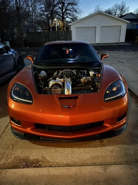 Front view of an orange Chevrolet Corvette with the hood open, revealing a modified engine with a large turbocharger.
