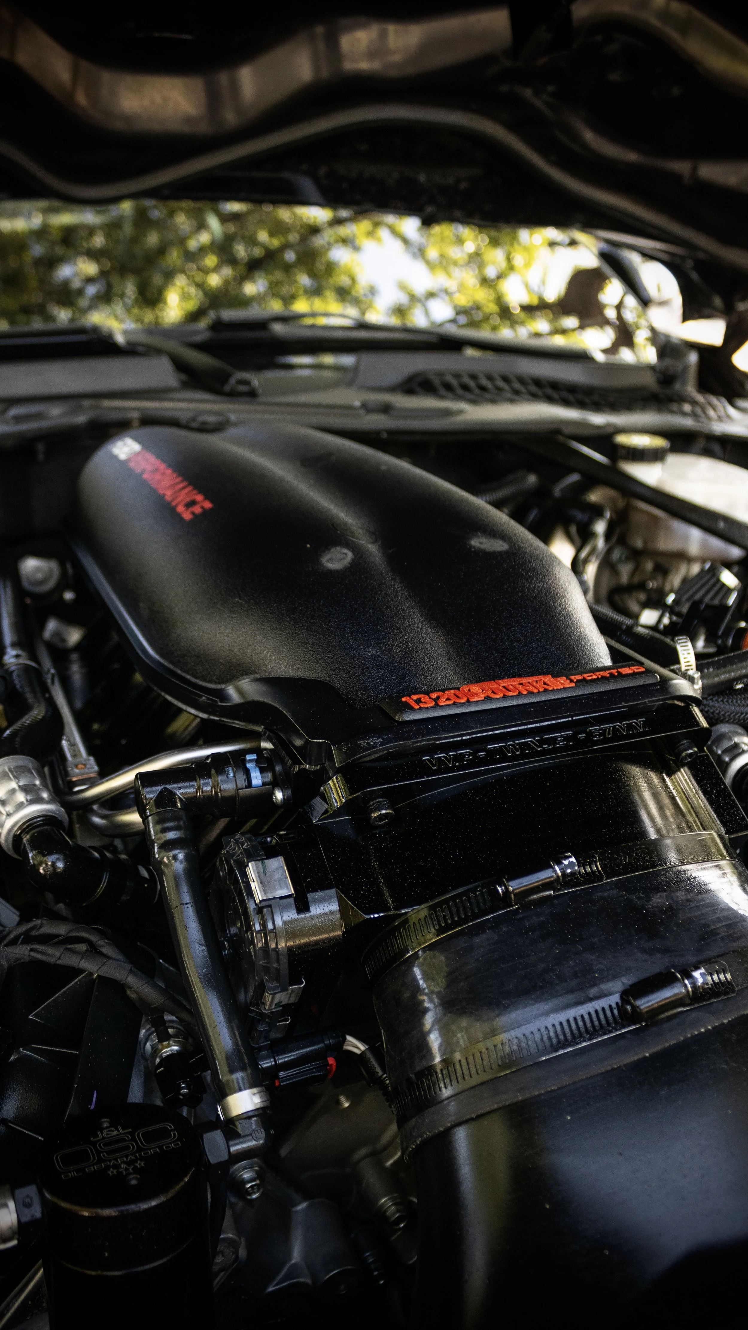 Close-up of a motorcycle engine with black components, some with red text, and various metallic parts, set outdoors with blurred trees in the background.