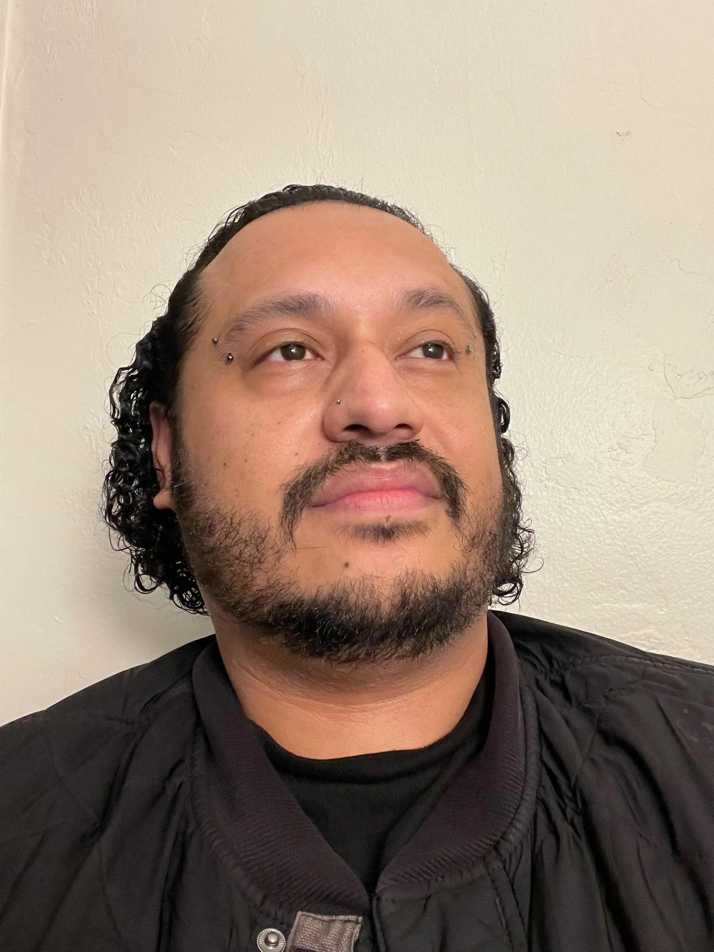 Close-up of a man with curly black hair and a beard, wearing a black jacket and shirt, standing against a plain off-white wall.