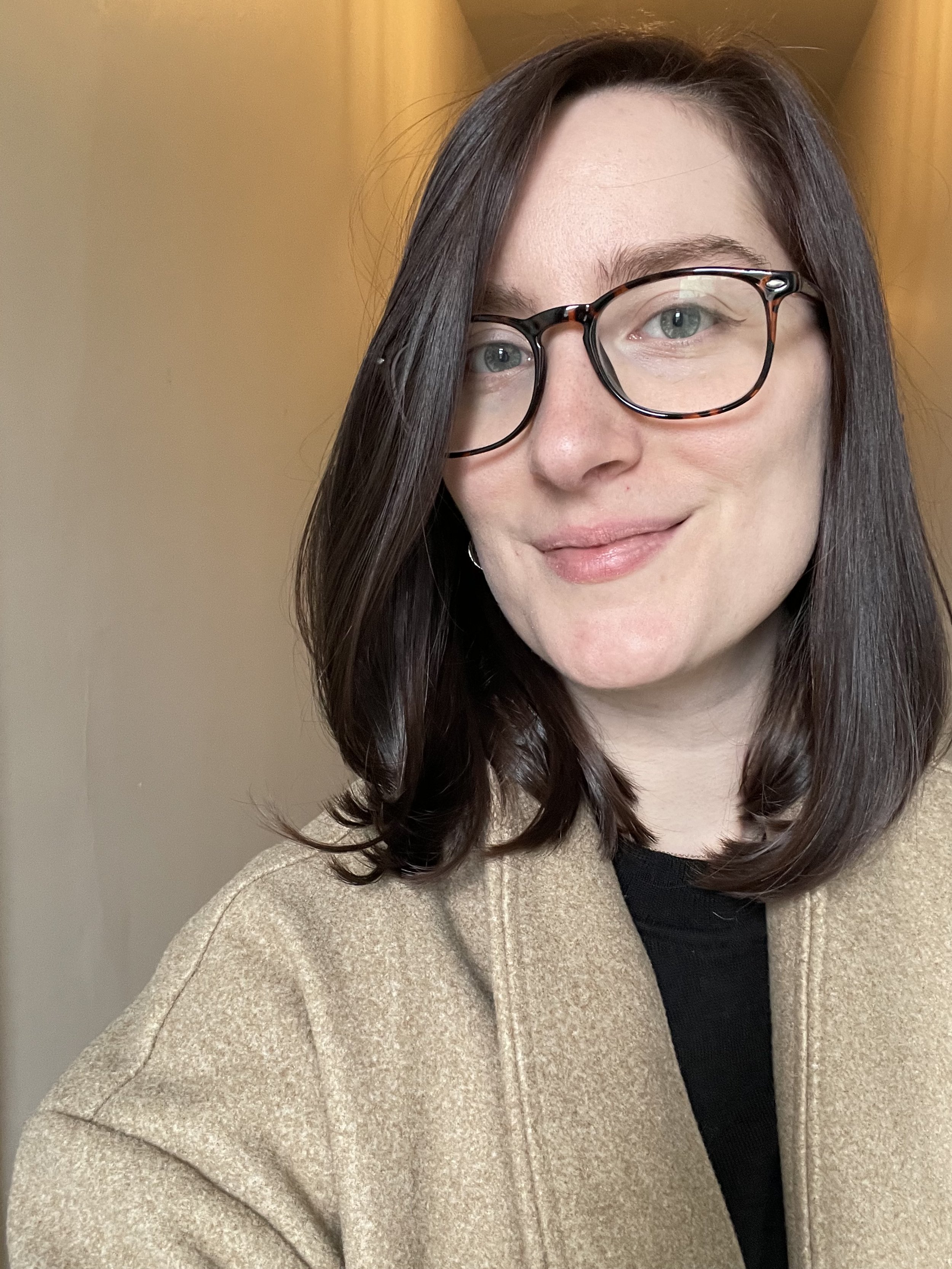 A woman with shoulder-length dark brown hair wearing black-rimmed glasses, a black top, and a tan coat, smiling softly indoors.