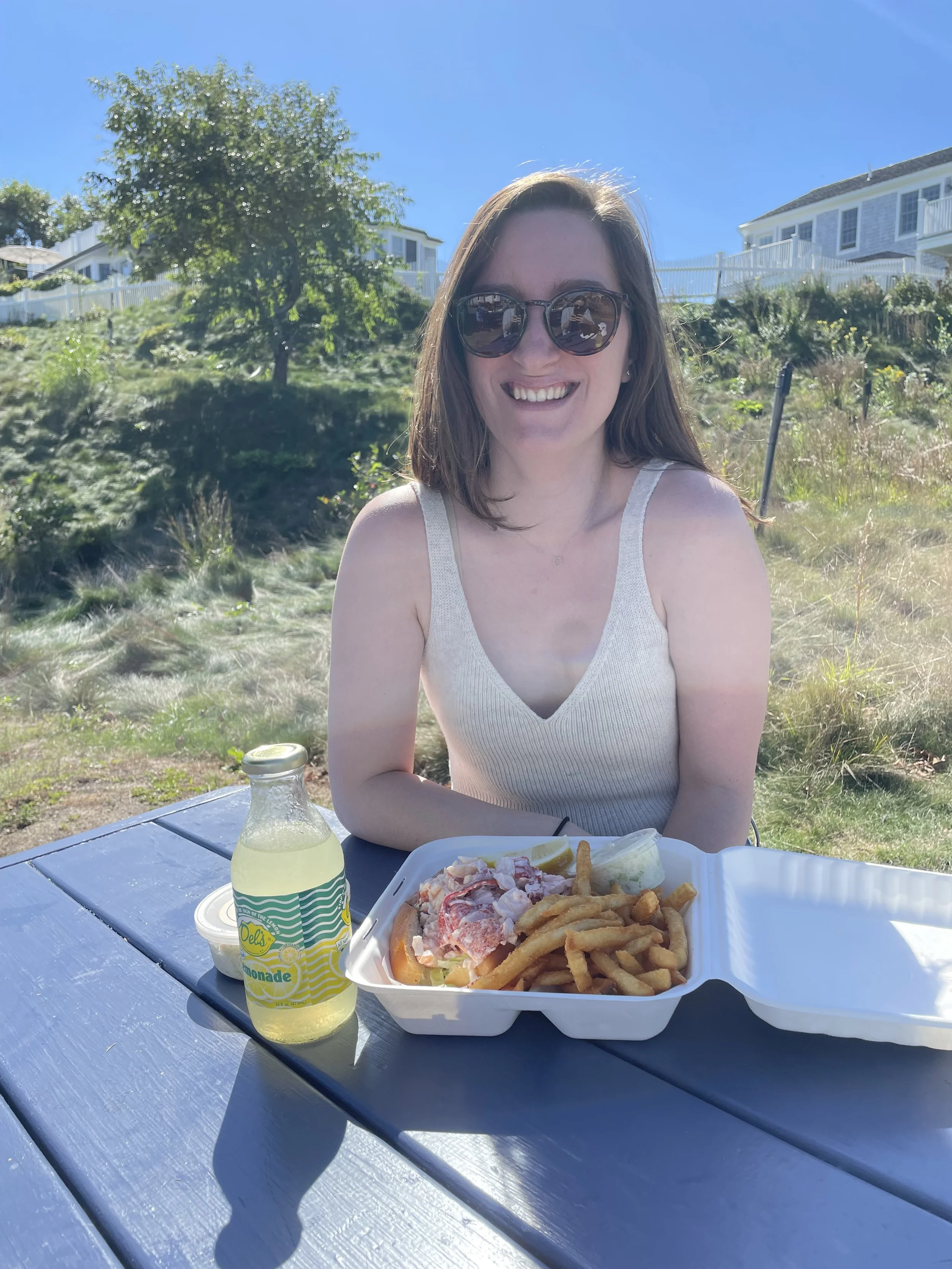 A woman wearing sunglasses and a sleeveless top is sitting at a blue picnic table outdoors, smiling, with a salad and French fries in a takeout container and a lemonade bottle nearby. The background features trees and houses under a clear blue sky.