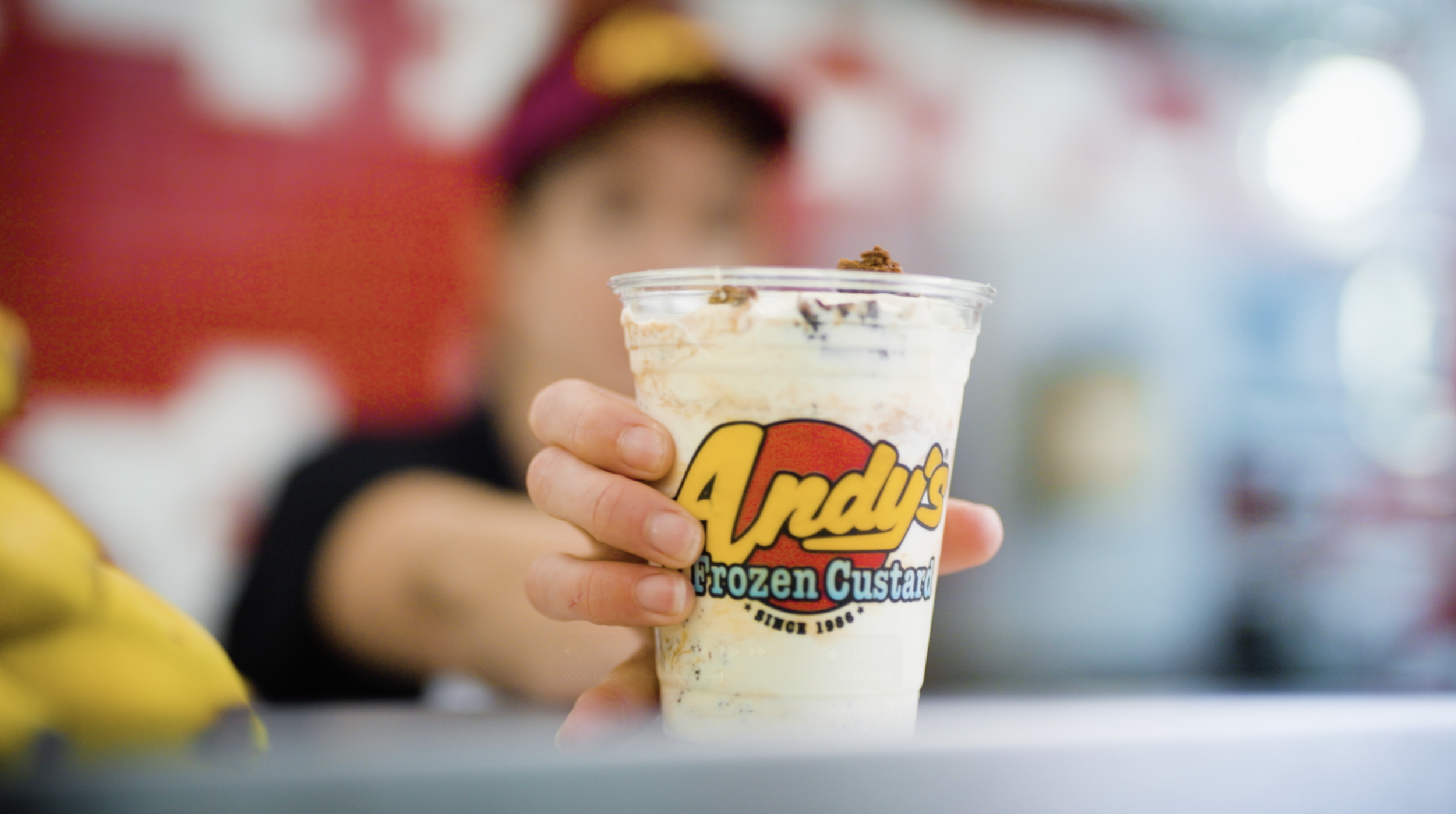 Close-up of a person's hand holding a cup of Andy's Frozen Custard with a blurred background of a person and store interior.