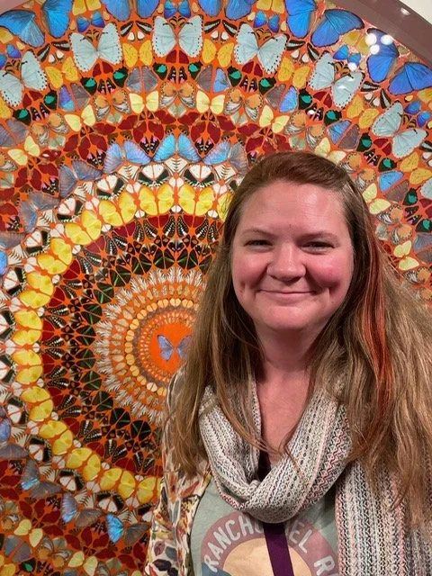 Massage therapist, esthetician, and owner of Big Hands Kind Heart studio standing in front of an image of brightly colored butterflies smiling.